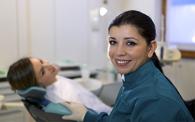 A closeup of a female dentist smiling with a female patient in the background laying in a dental chair